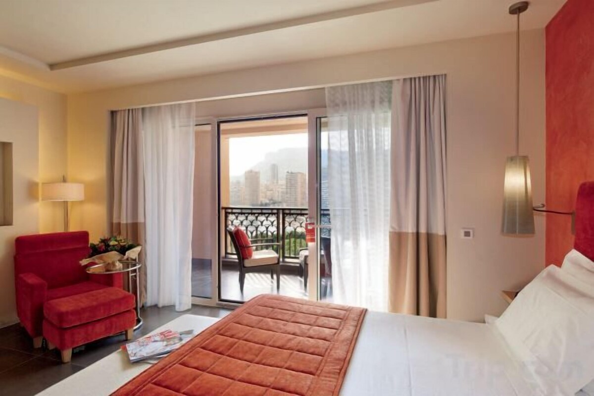 A hotel room with a bed, a red armchair, a small table with a lamp, and a balcony with a chair.
