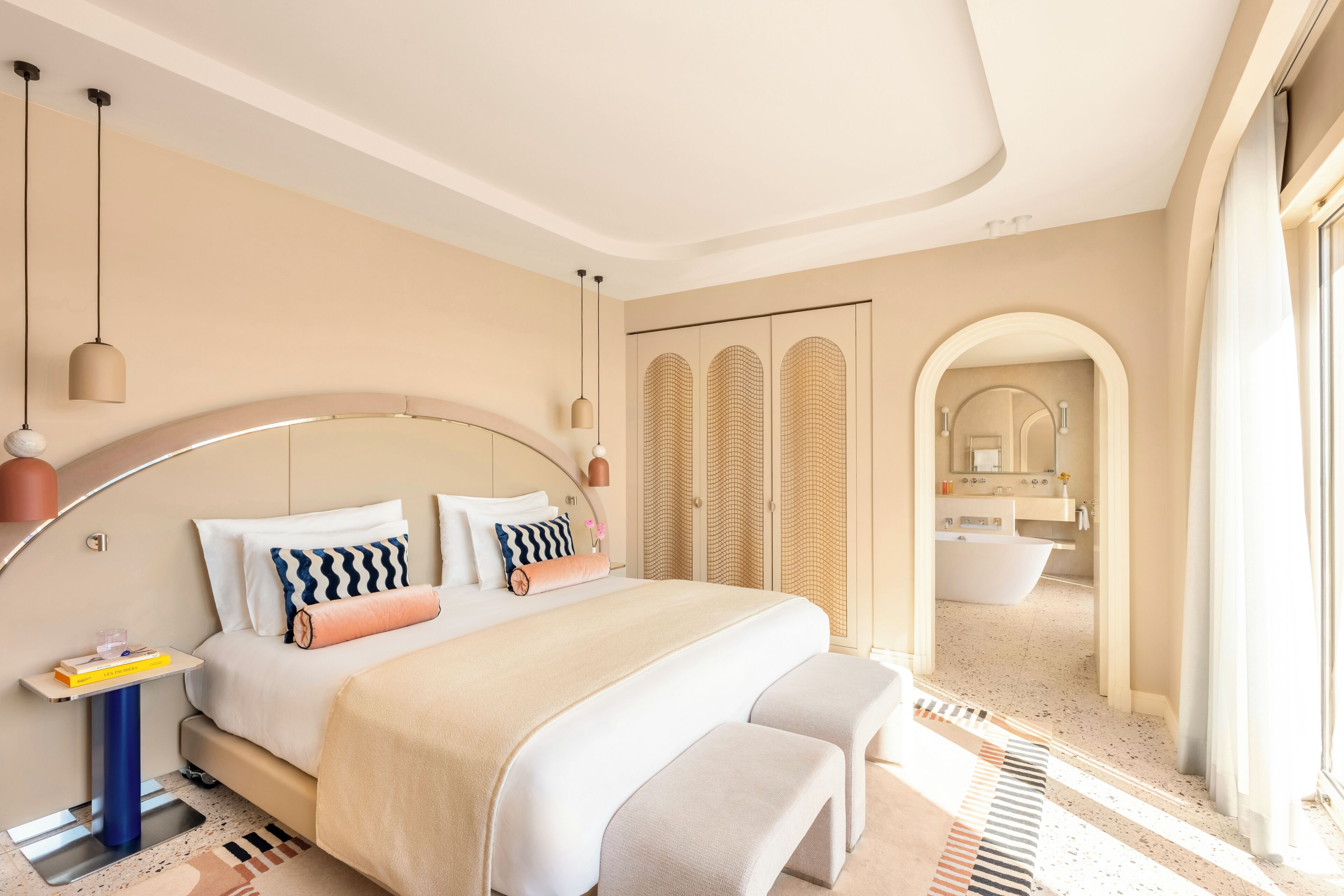 Un dormitorio moderno con un cabecero curvo, una cama grande con muchas almohadas, mesitas de noche y vistas al baño.