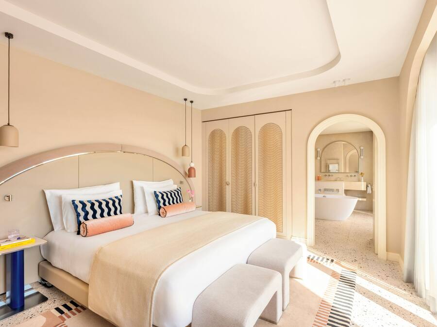 Un dormitorio moderno con un cabecero curvo, una cama grande con muchas almohadas, mesitas de noche y vistas al baño.