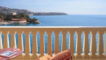 Un balcon avec vue sur la mer, une chaise, une table avec un livre et un verre d’eau.
