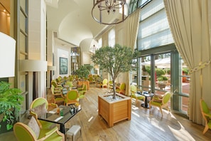 A spacious indoor area with multiple seating arrangements, a central tree planter, and large windows providing natural light. - Monte-Carlo Bay Hotel & Resort (Monaco)