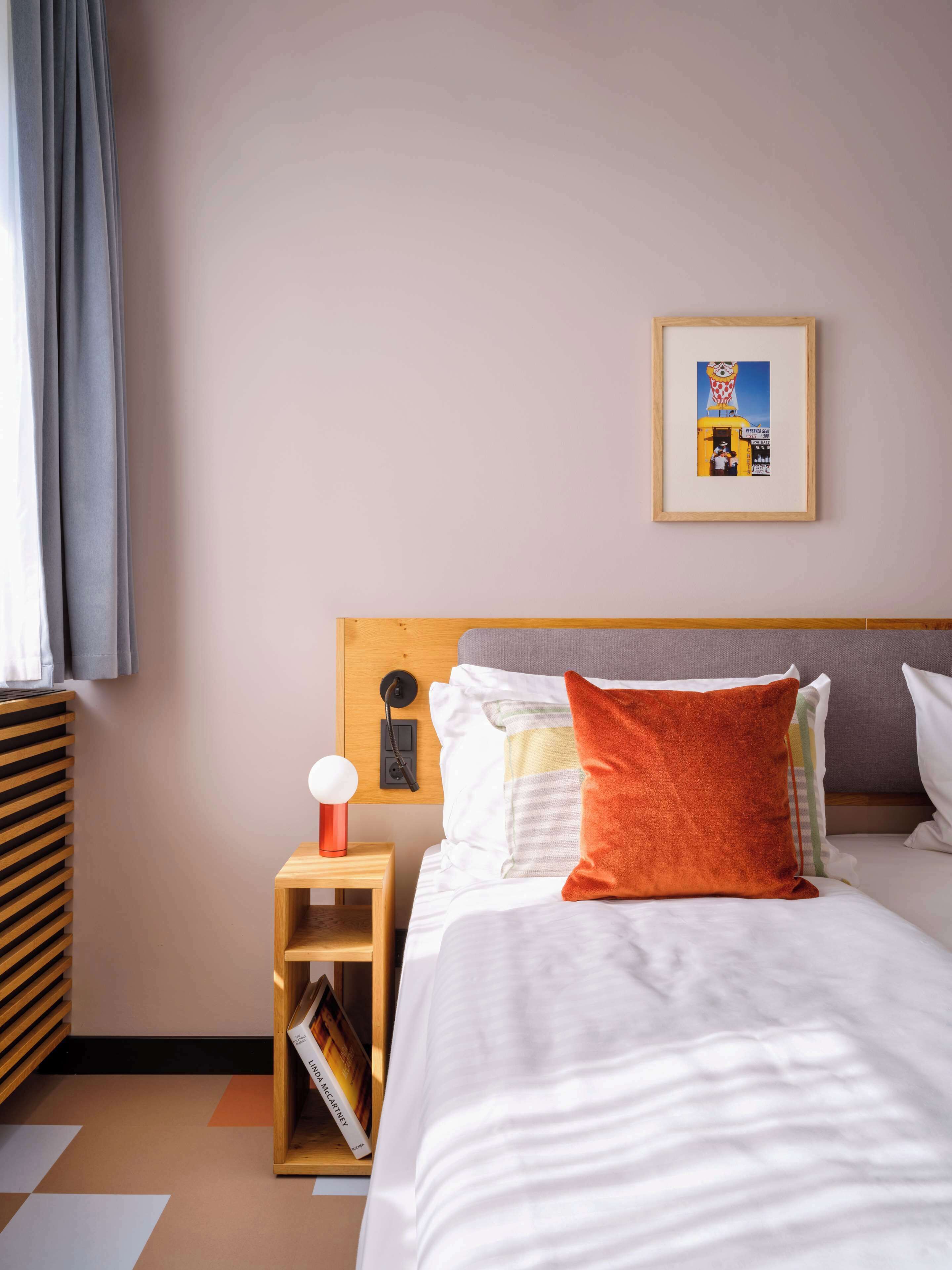 A hotel room with a bed, a nightstand, a bedside table, a framed picture on the wall, and a window with curtains.