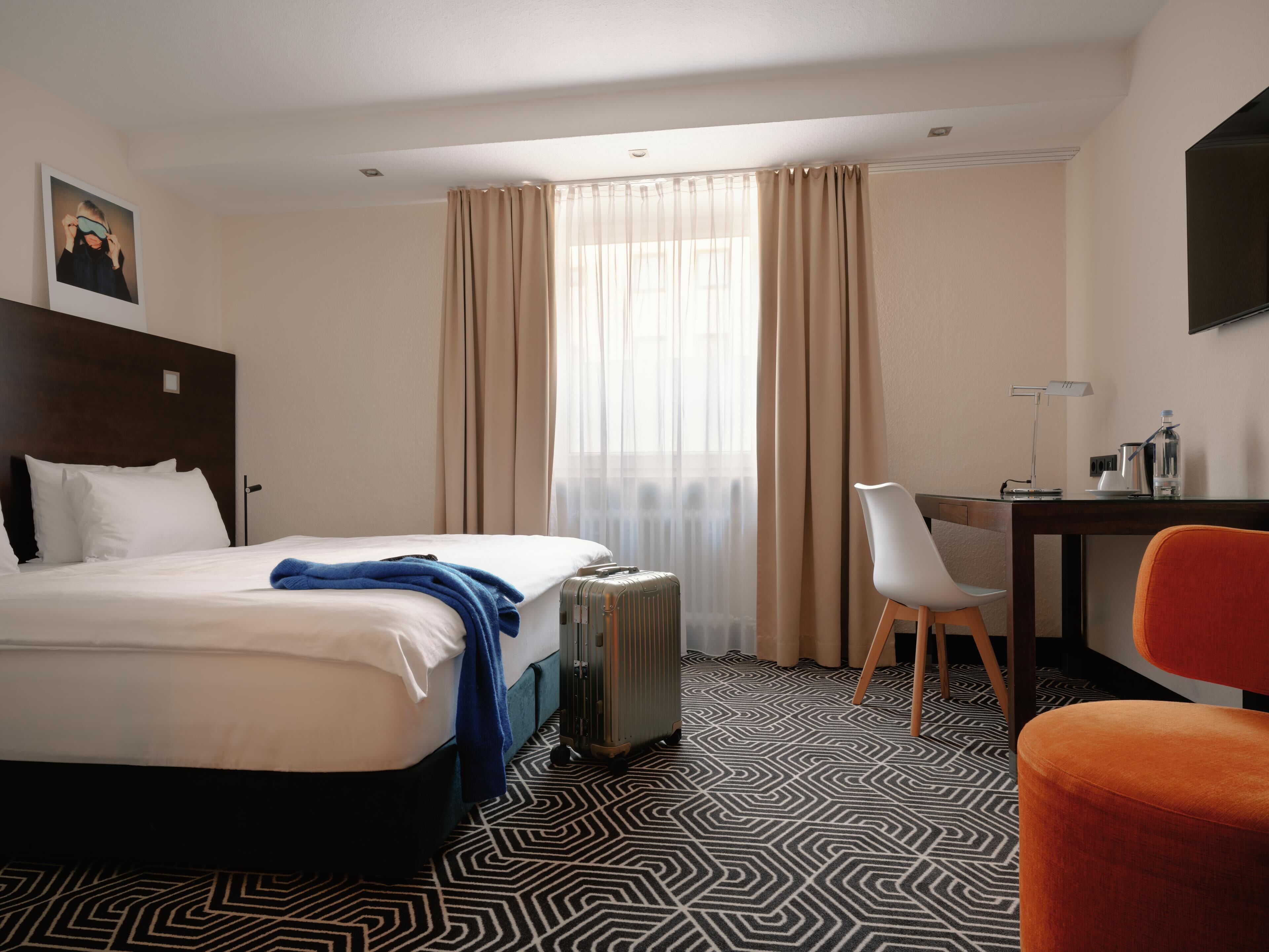 Una habitación de hotel con cama, escritorio, silla, maleta y radiador.