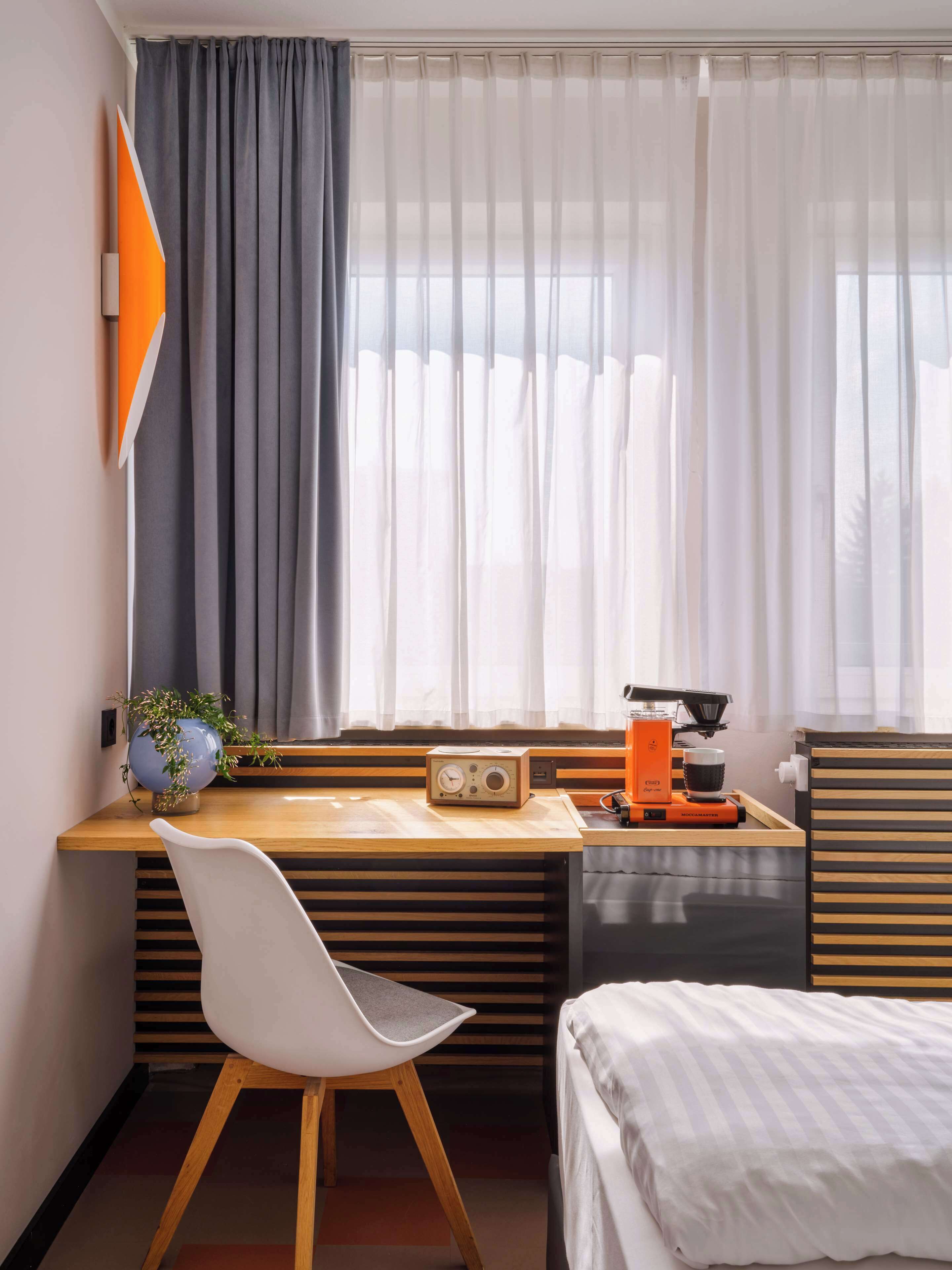 Une chambre d’hôtel moderne avec un bureau en bois, une chaise, un lit et une fenêtre avec des rideaux légers.