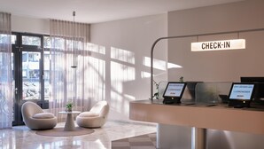 A modern reception area with a check-in counter, two armchairs, a small table with a plant, and a large window allowing natural light. - Flemings Hotel München Schwabing (Munich)