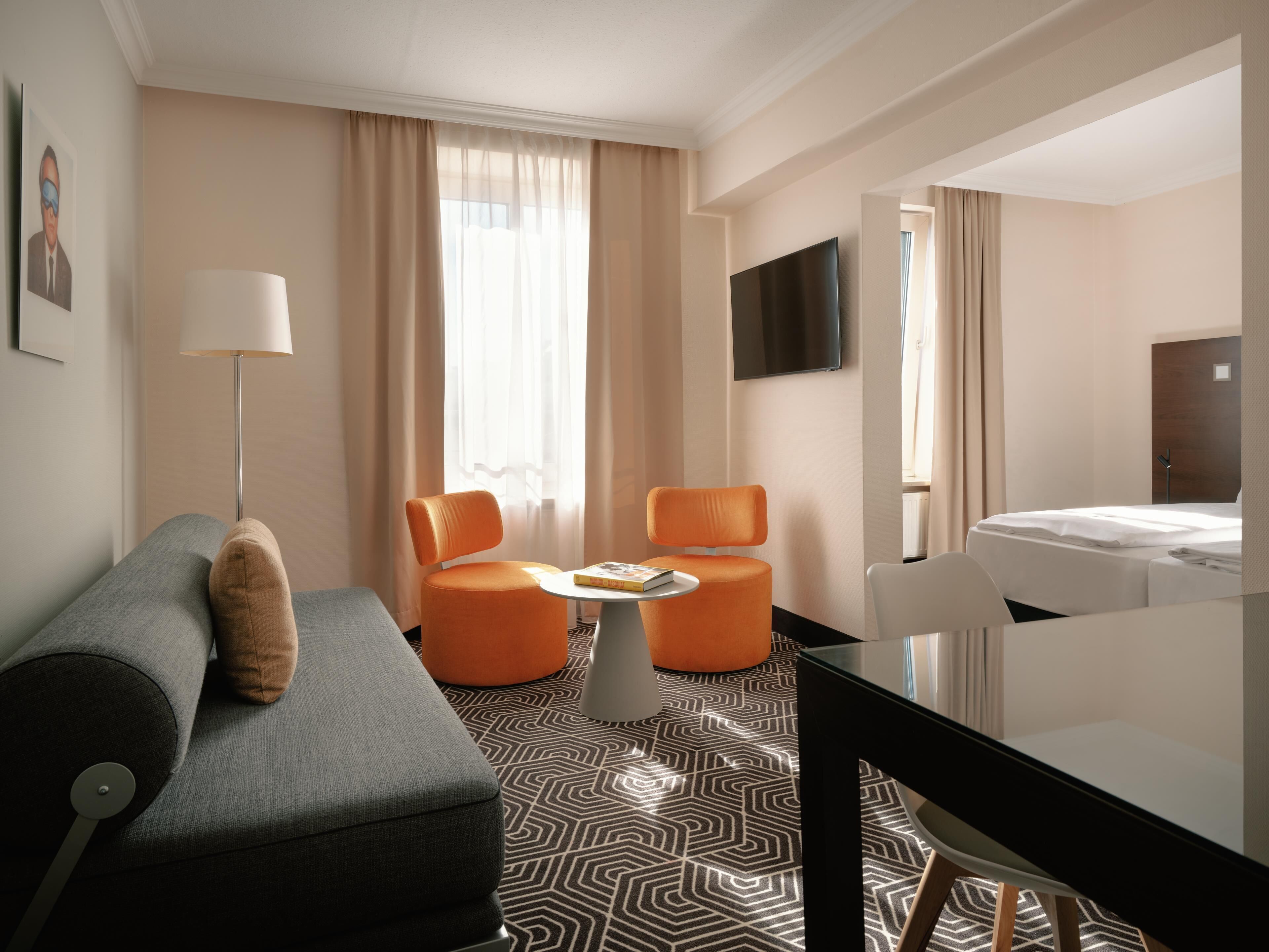 Hotelkamer met een grijze bank, oranje fauteuils, een klein rond tafeltje, een aan de muur bevestigde televisie en een bed met witte lakens.