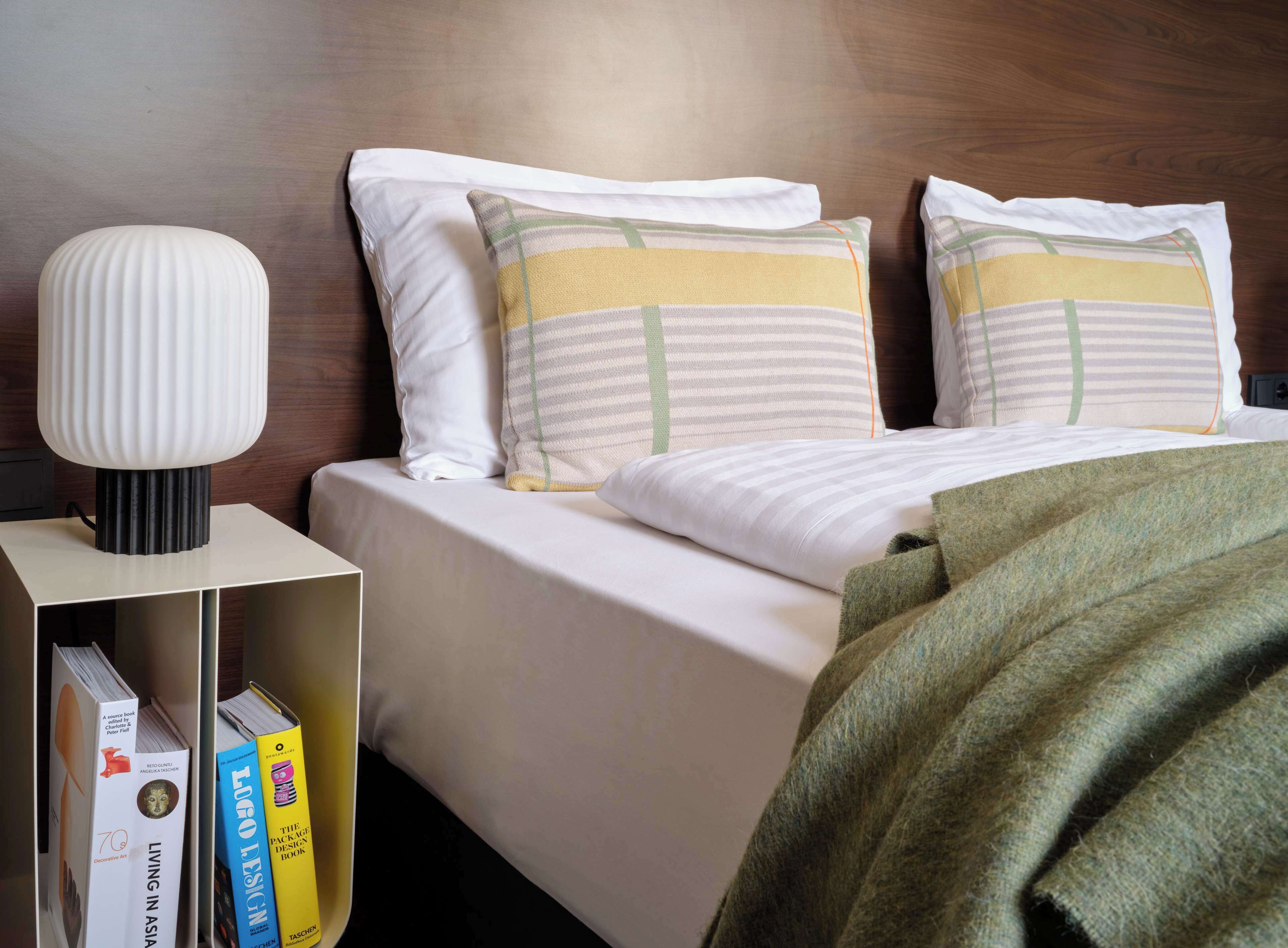 Una cama bien hecha con sábanas blancas, dos almohadas con diseños a rayas, una lámpara de noche y una estantería con libros.