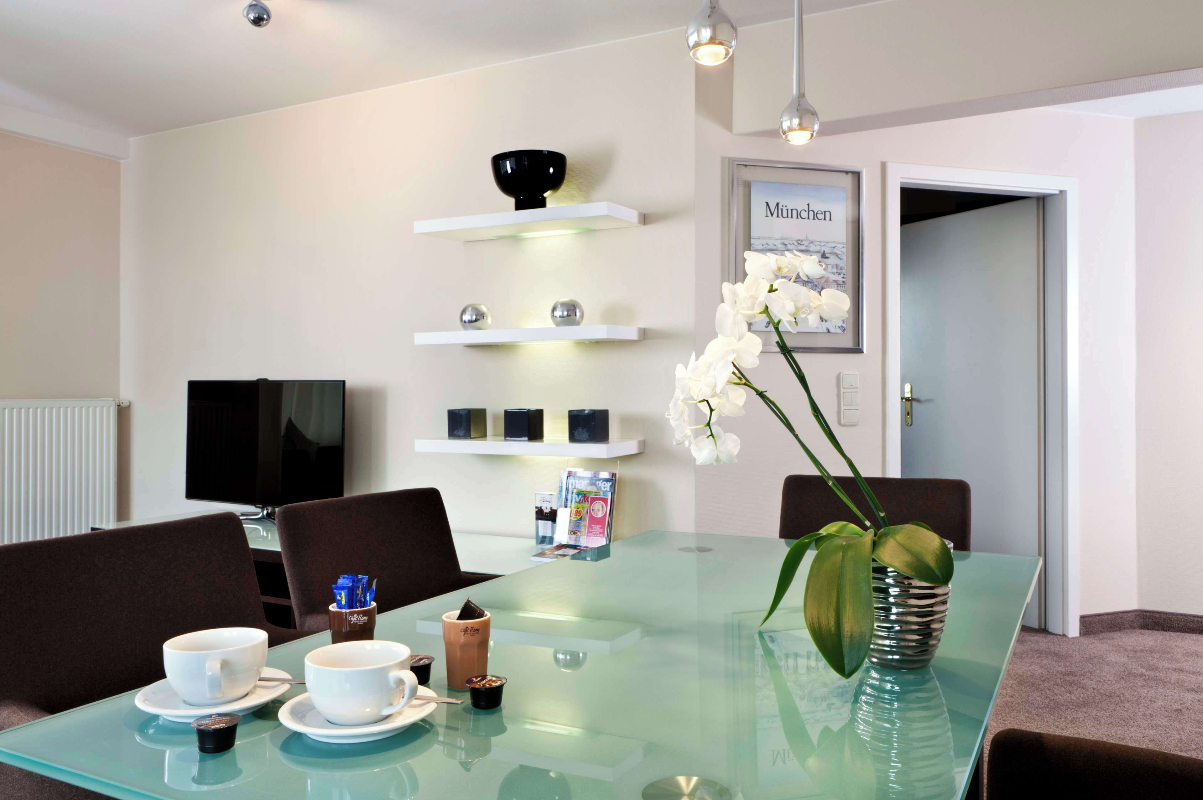 Un comedor moderno con una mesa de vidrio, tazas blancas, un jarrón con orquídeas y un cuadro en la pared.