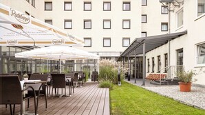 An outdoor patio with wicker furniture and umbrellas, a grassy area, and a building with multiple windows. - Flemings Hotel München Schwabing (Munich)