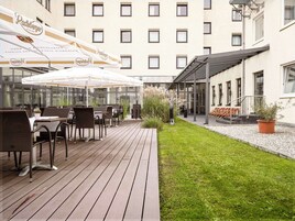 Un patio exterior con mobiliario de mimbre y sombrillas, una zona con césped y un edificio con múltiples ventanas.