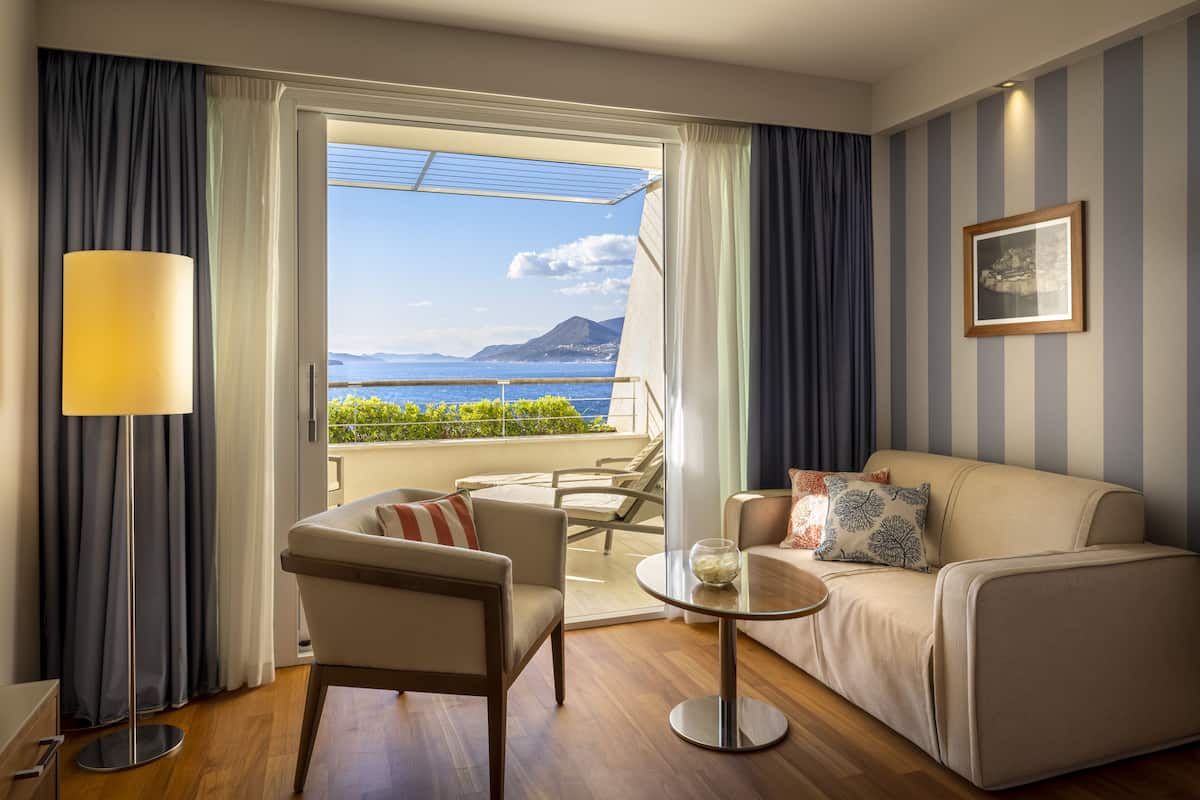 A hotel room with a balcony, a sofa, a chair, a table, and a lamp.