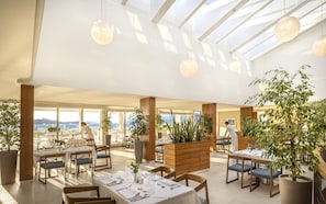 A spacious restaurant with high ceilings, large windows, and a view of the sea. Tables are set with white tablecloths and silverware.