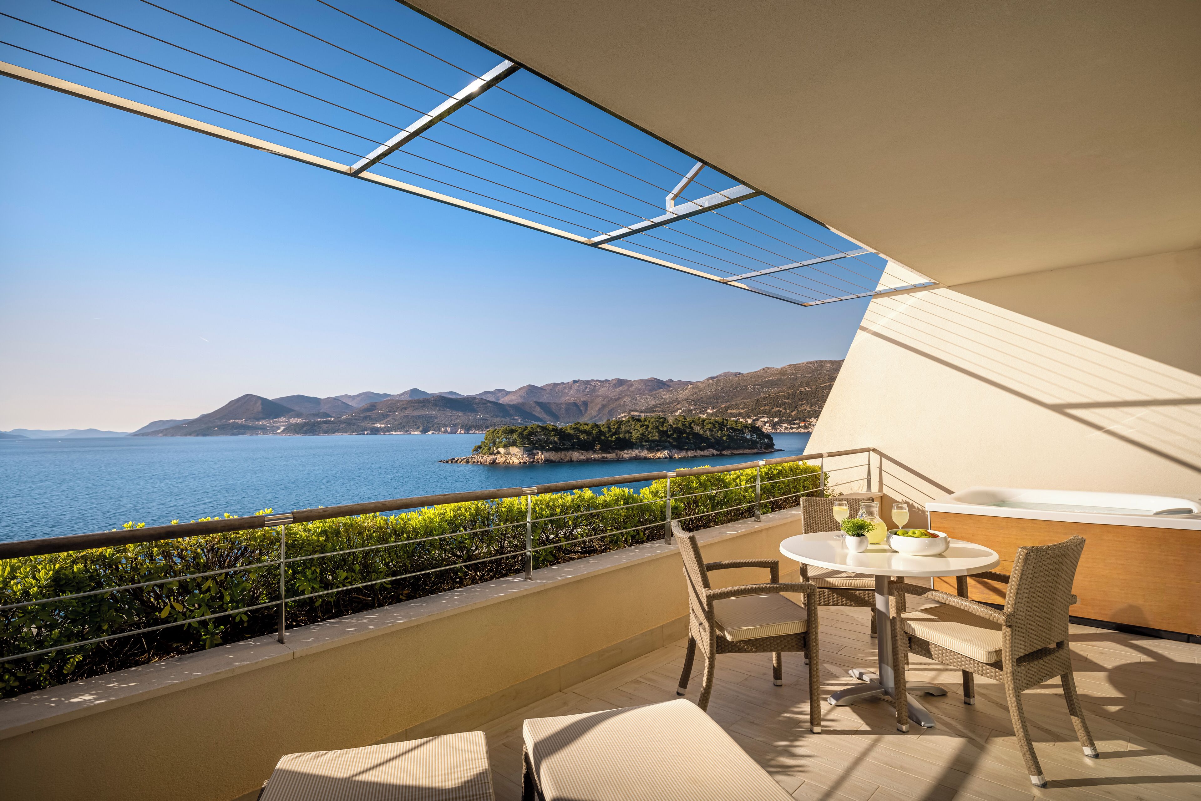 A balcony with a view of the sea and mountains, a round table with chairs, and a small garden area.