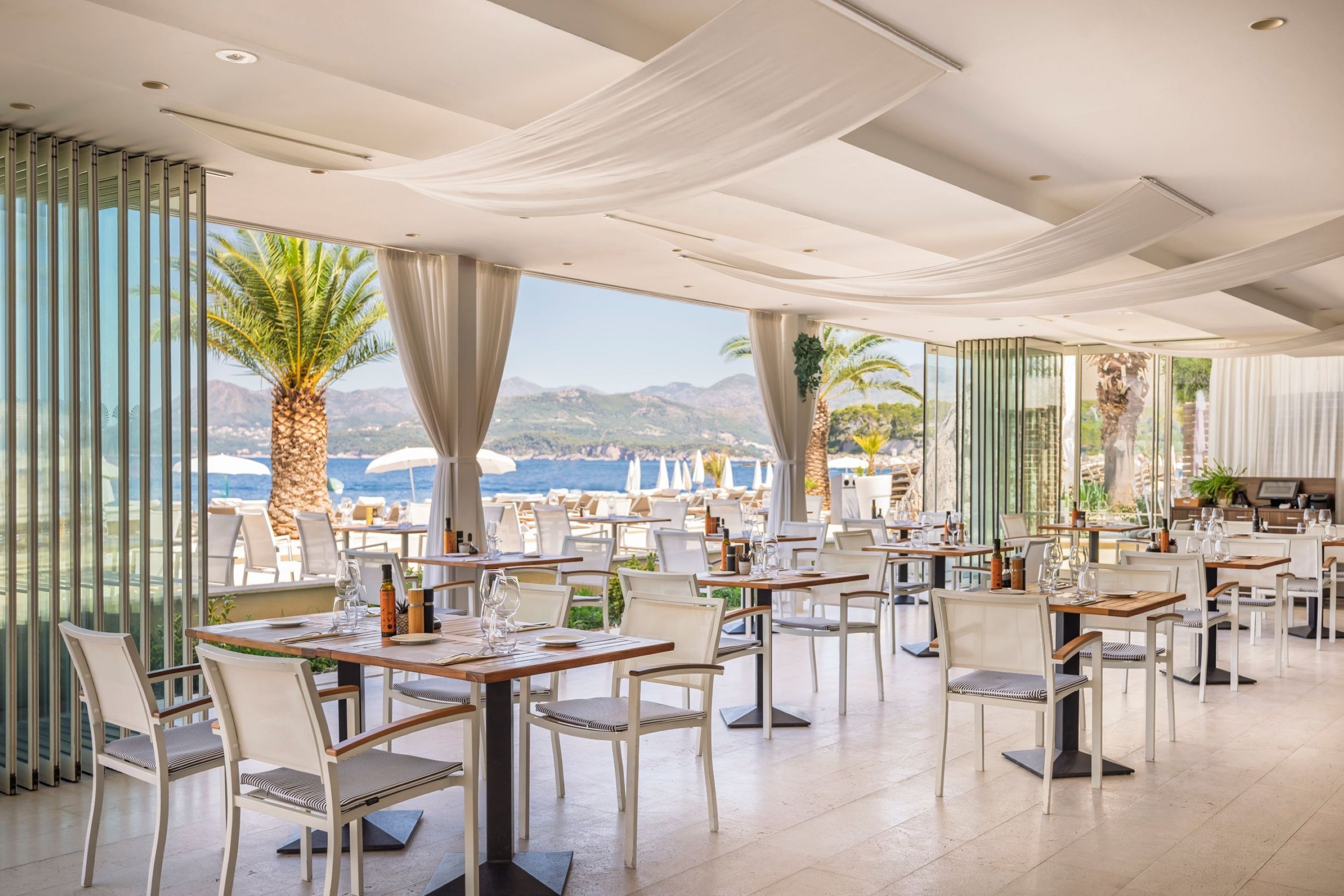 Un restaurant avec des tables et des chaises, de grandes fenêtres et une vue sur la mer et les montagnes.