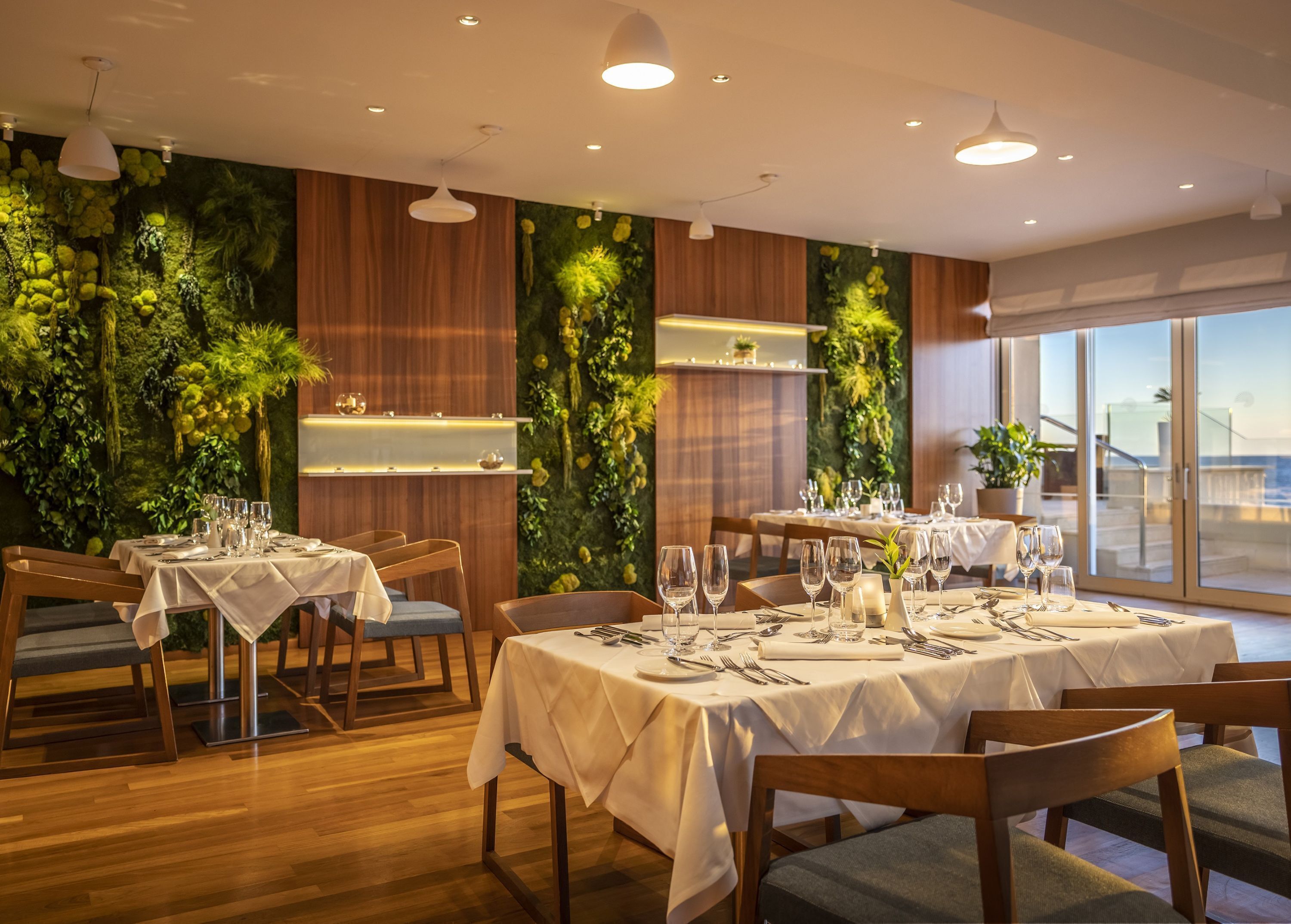 Um restaurante com mesas e cadeiras de madeira, uma parede verde com plantas e vista para o mar.