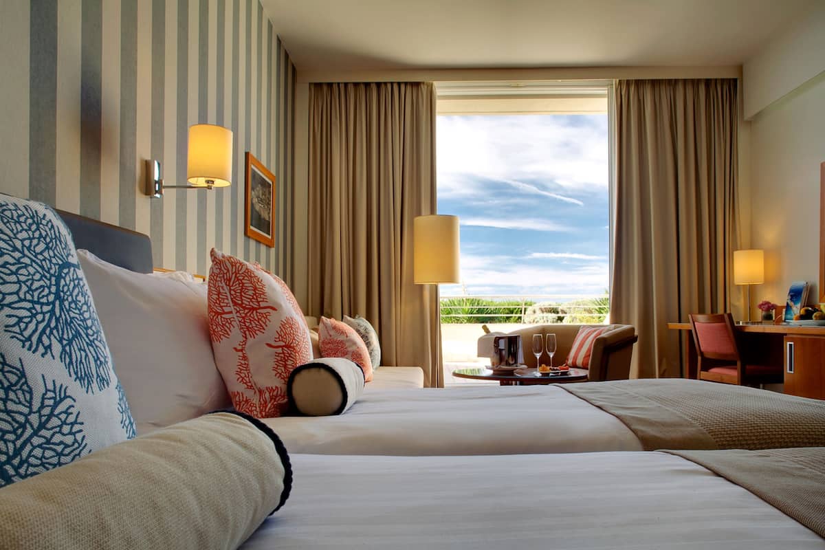 A hotel room with two beds, a desk, a chair, and a view of the outdoors.