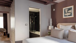 Une chambre à coucher avec un grand lit, une table de chevet, une commode, une lampe fixée au mur, un tableau encadré et une porte donnant sur une salle de bain avec douche.