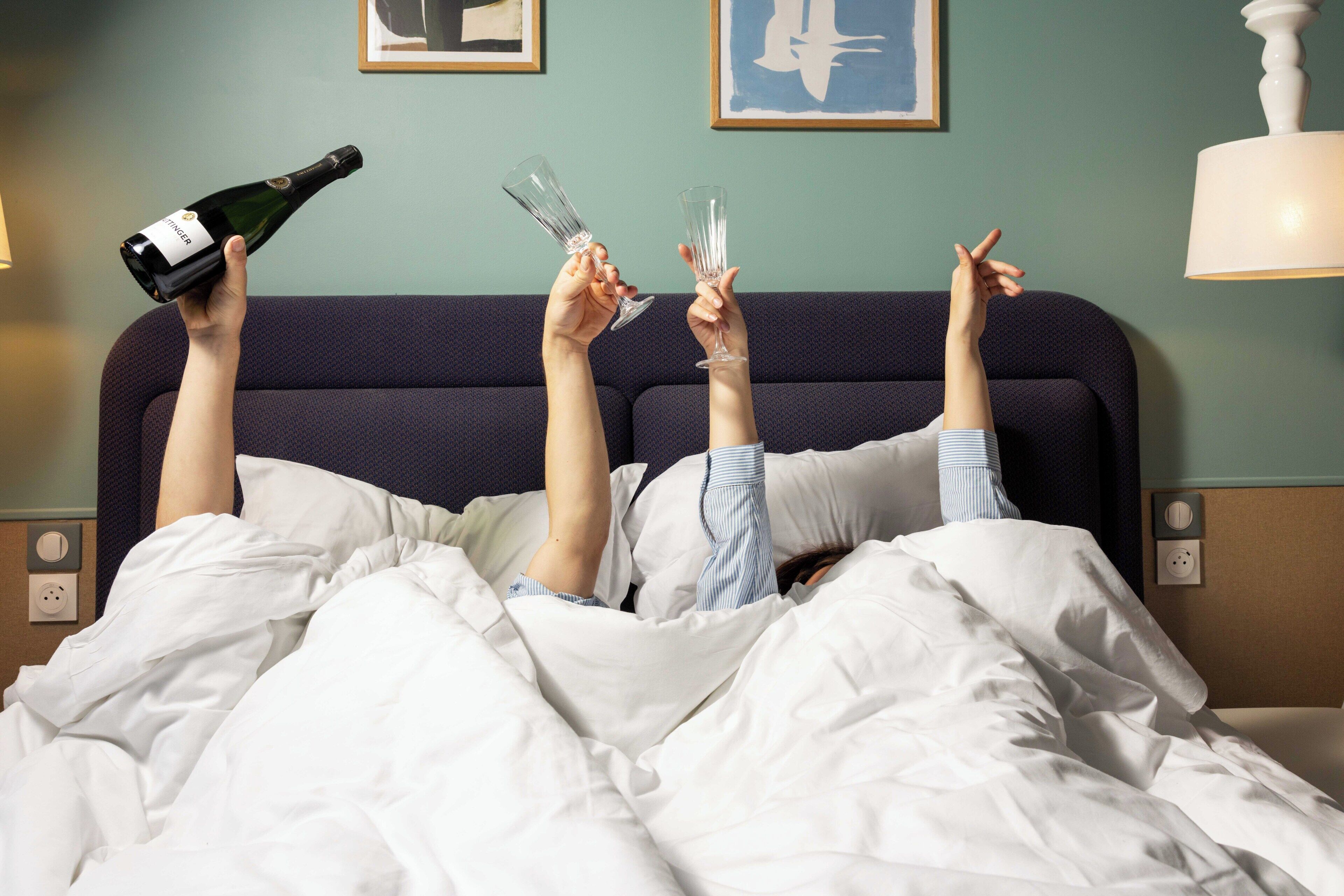 Une personne allongée dans un lit, avec des coupes à champagne et une bouteille à proximité.