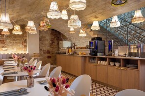 A restaurant with multiple dining tables, hanging floral chandeliers, a brick wall, and a kitchen area with modern appliances. - Hôtel MARAIS HÔme (Paris)