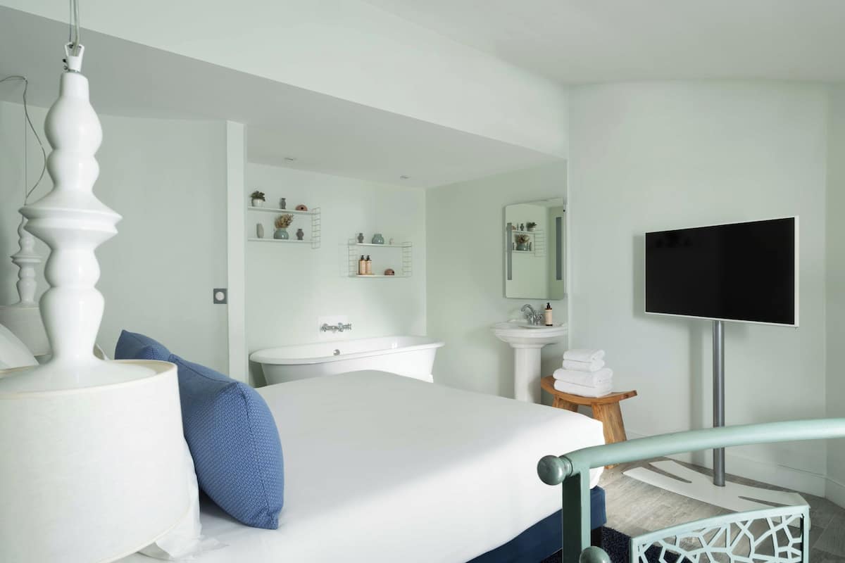 A bedroom with a bed, a bedside lamp, a TV, a bathroom with a sink and mirror, and a small wooden stool.