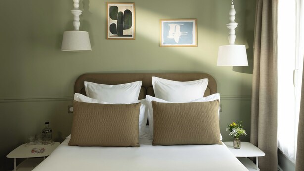 Habitación de hotel con cama, dos almohadas, mesita de noche, un jarrón con flores y dos obras de arte en la pared.