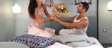 A couple on a bed with a bouquet of flowers, a round mirror, and a shelf with decorative items.