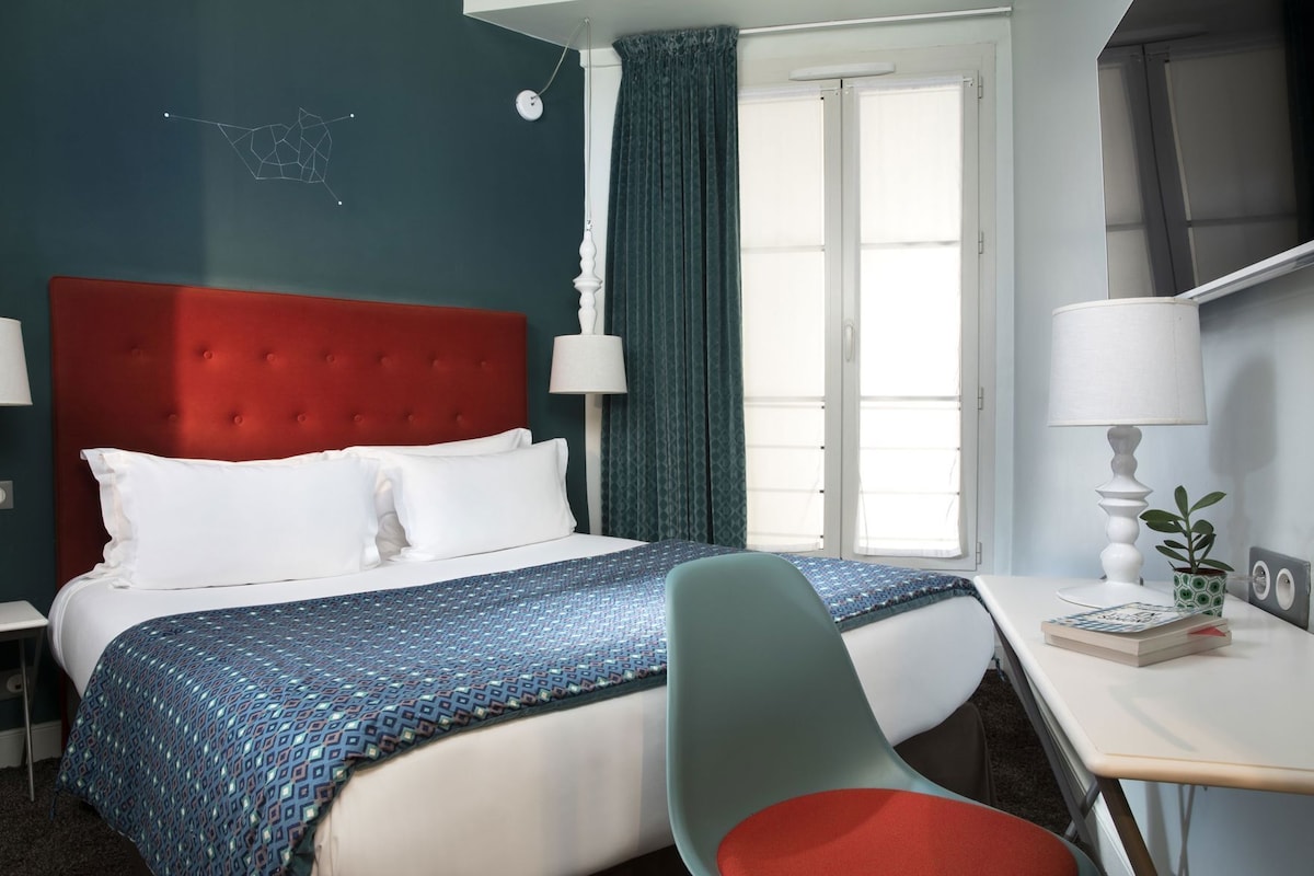 Una camera d'albergo con un letto, una scrivania, una sedia, una lampada, una pianta in vaso e una parete con motivo geometrico.