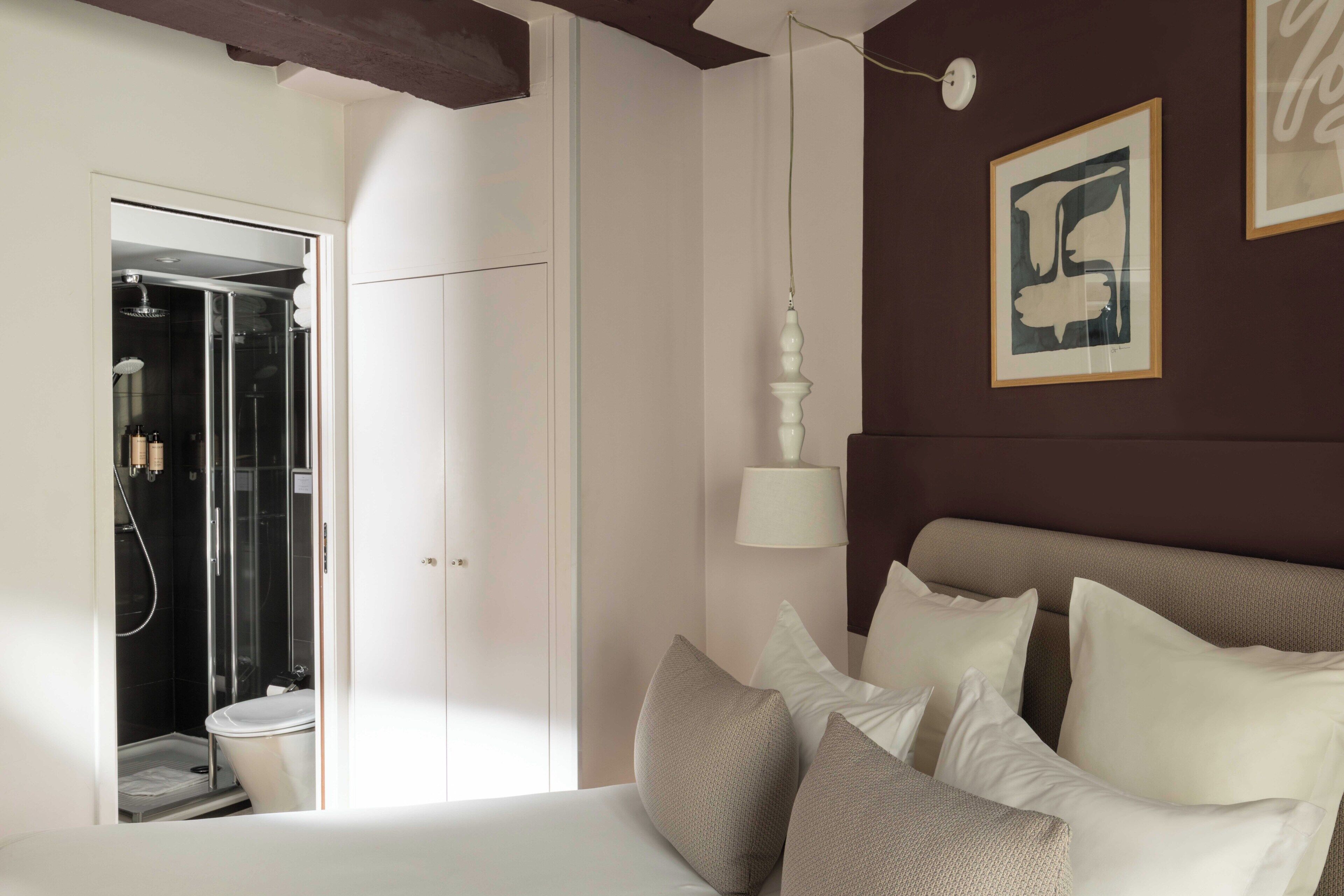A bedroom with a bed, pillows, a nightstand, a wall-mounted lamp, and a framed picture.