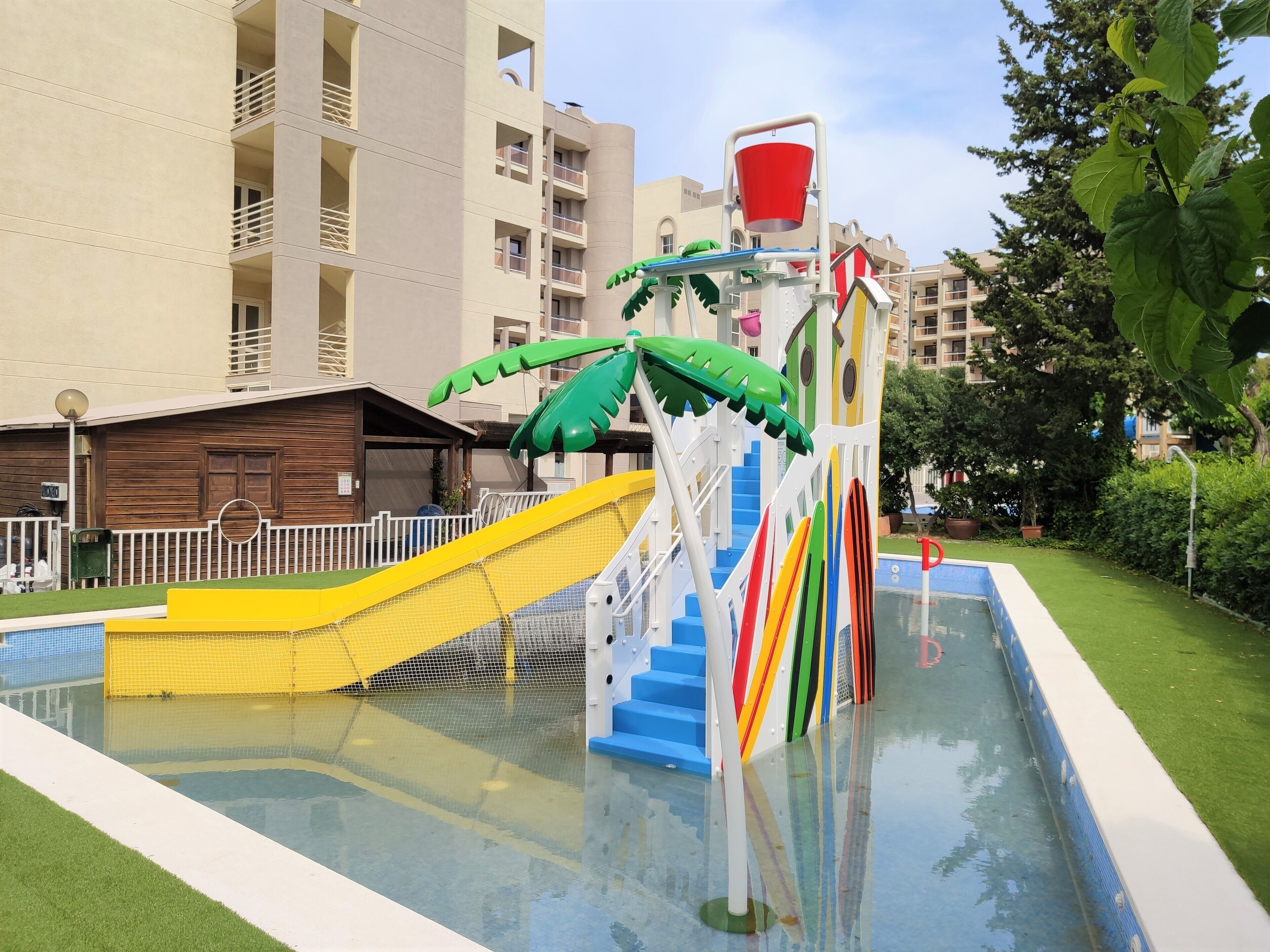 A water play area with slides and a pool, surrounded by a fence and greenery.