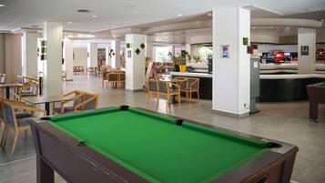 A pool table in a modern indoor setting with wooden chairs and tables, a bar area, and a seating area with a sofa.