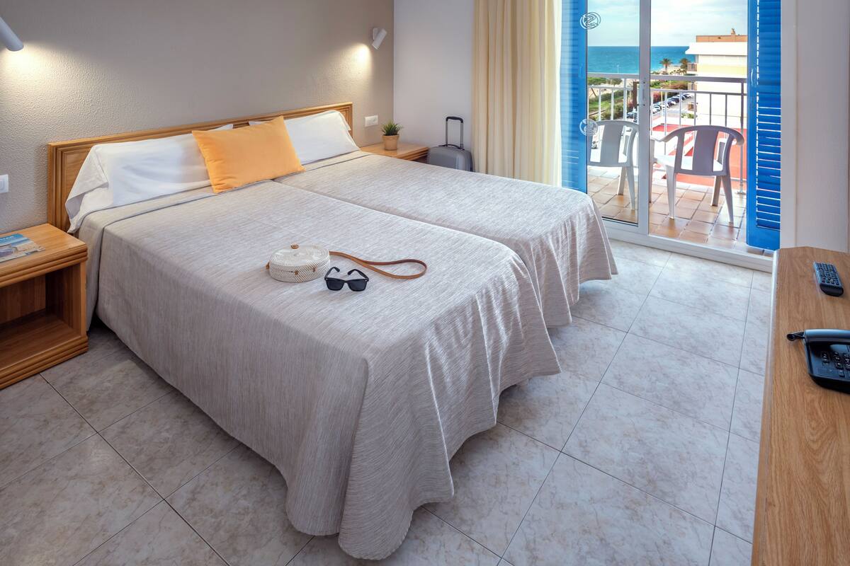 Habitación de hotel con dos camas, un balcón con sillas y vista al mar.