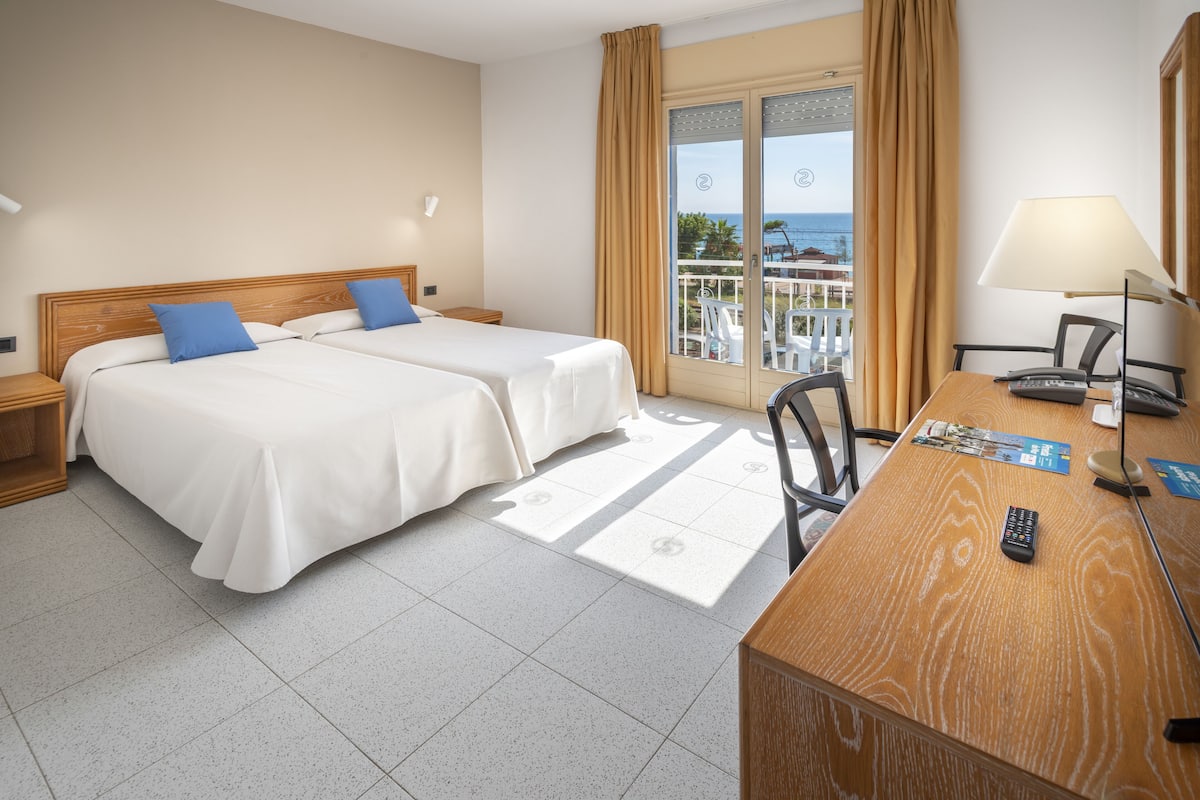 Habitación de hotel con dos camas, un escritorio con lámpara, una silla y vista al mar a través de un ventanal grande.