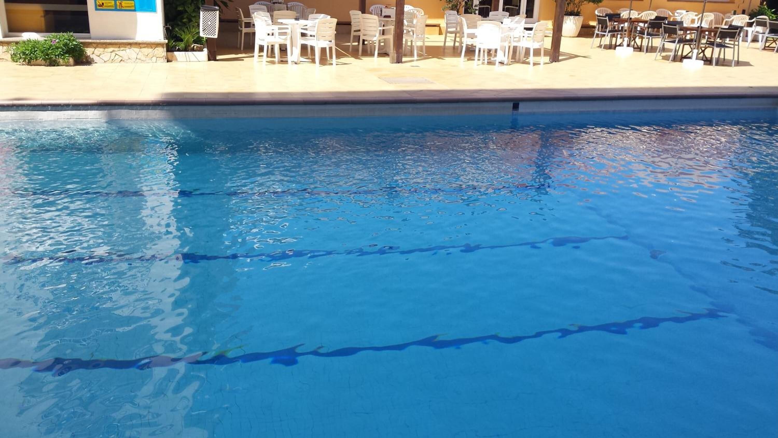 En swimmingpool med klart, blåt vand og linedelere. Hvide plastikstole og borde er placeret rundt om poolområdet.