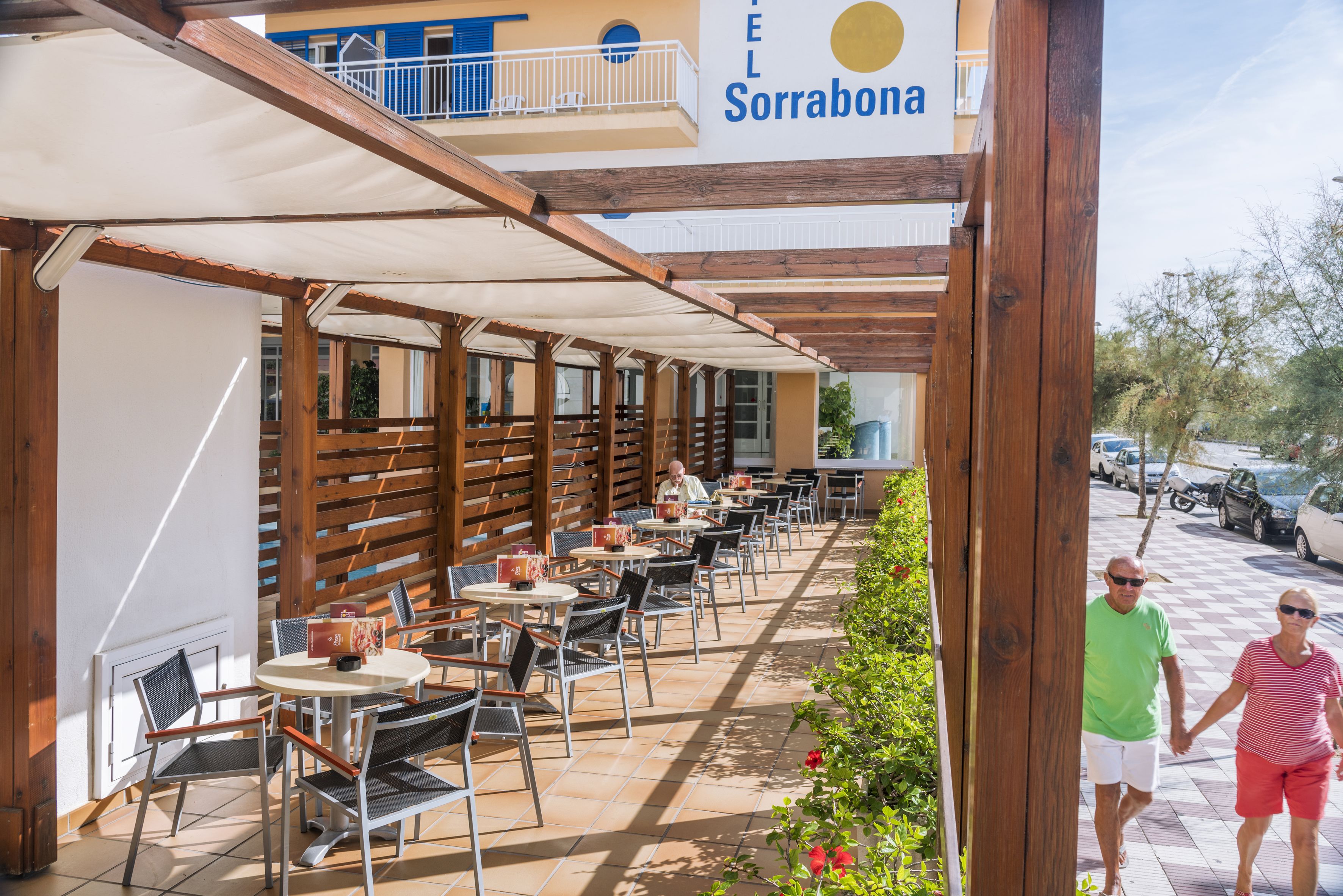 Un café al aire libre con mesas y sillas, una pérgola de madera y un letrero que dice "TEL Sorrabona".