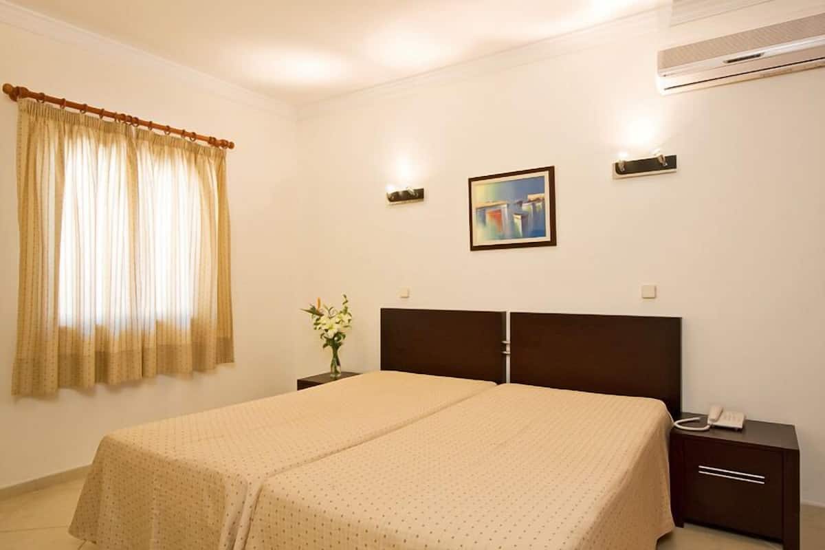 A hotel room with a double bed, a nightstand with a phone, a painting on the wall, and an air conditioning unit.