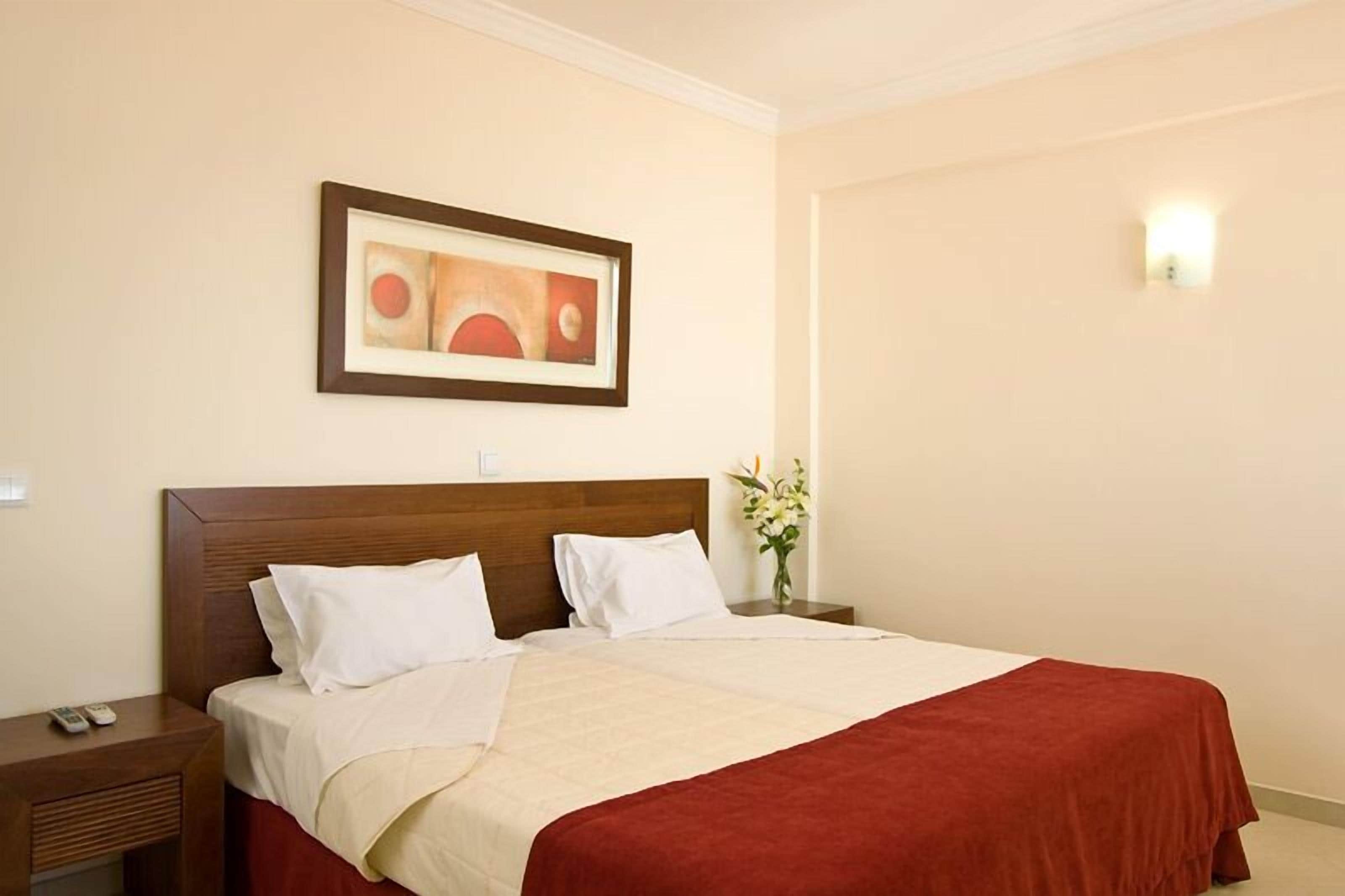A hotel room with a large bed, a nightstand, a framed picture on the wall, and a vase with flowers.