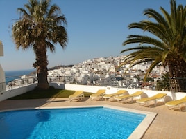 Una piscina con tumbonas, palmeras y vistas a un pueblo costero.