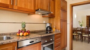 A kitchen with wooden cabinets, a granite countertop, stainless steel appliances, and a dining area visible through an open doorway. - Apartamentos Silmar (Albufeira)
