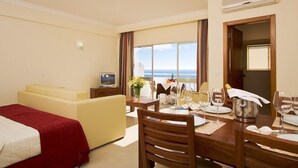 A hotel room with a bed, a sofa, a dining table set for two, and a large window offering a view of the sea. - Apartamentos Silmar (Albufeira)