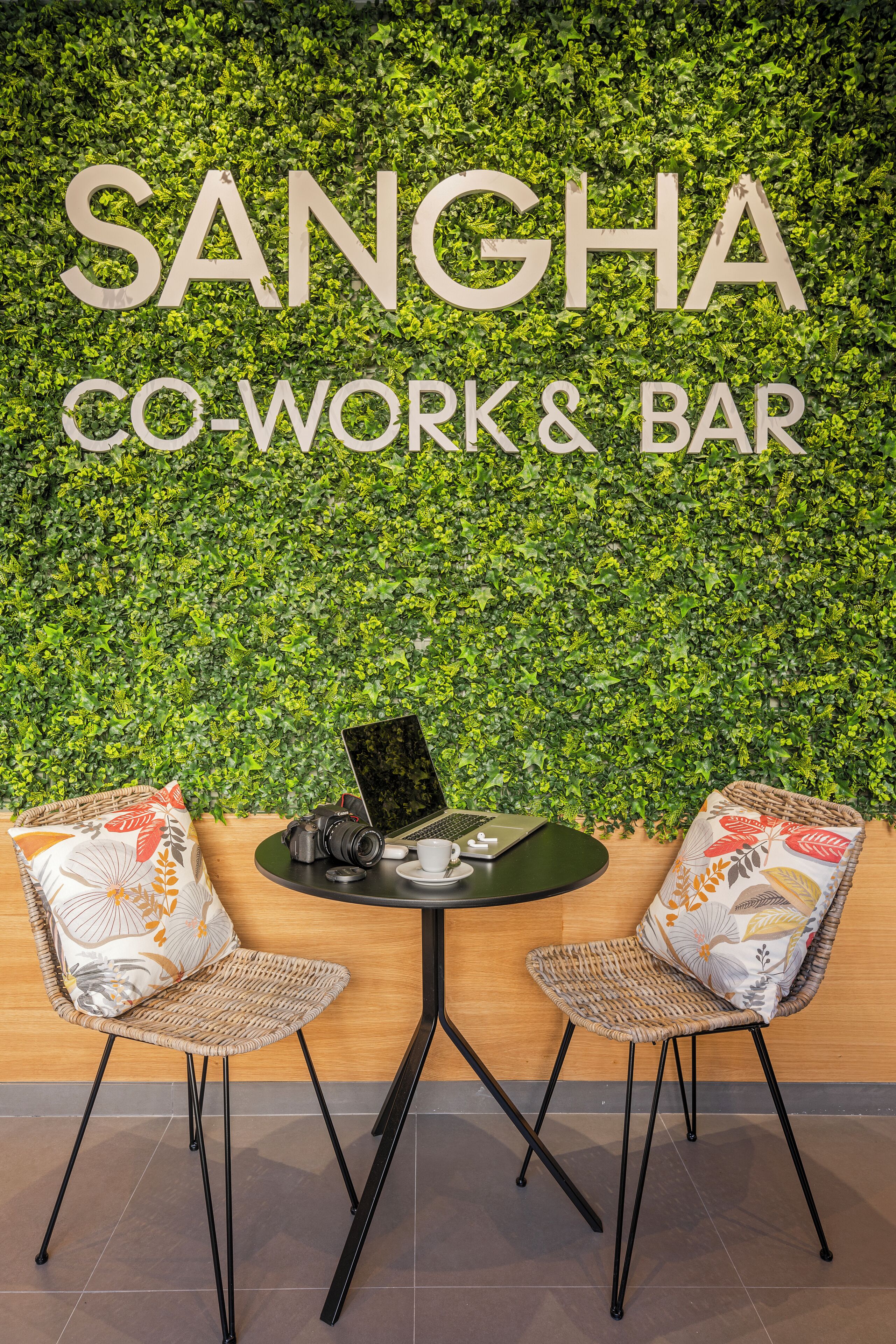 Un espacio de co-trabajo con una pared verde, dos sillas de mimbre, una laptop, una cámara y una taza.
