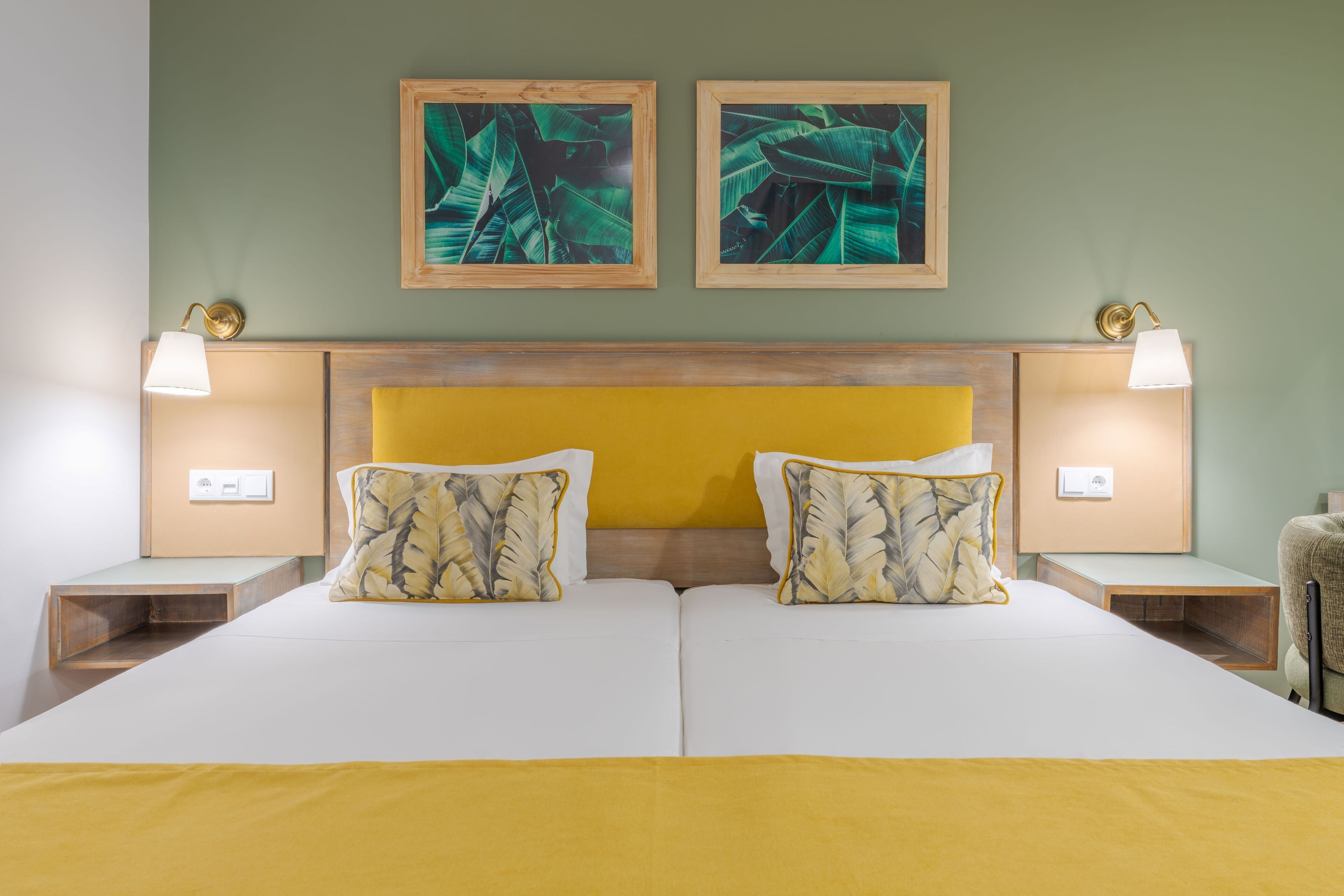 Una cama doble con cabecera de madera, dos mesitas de noche con lámparas y dos láminas enmarcadas con motivos botánicos en la pared.