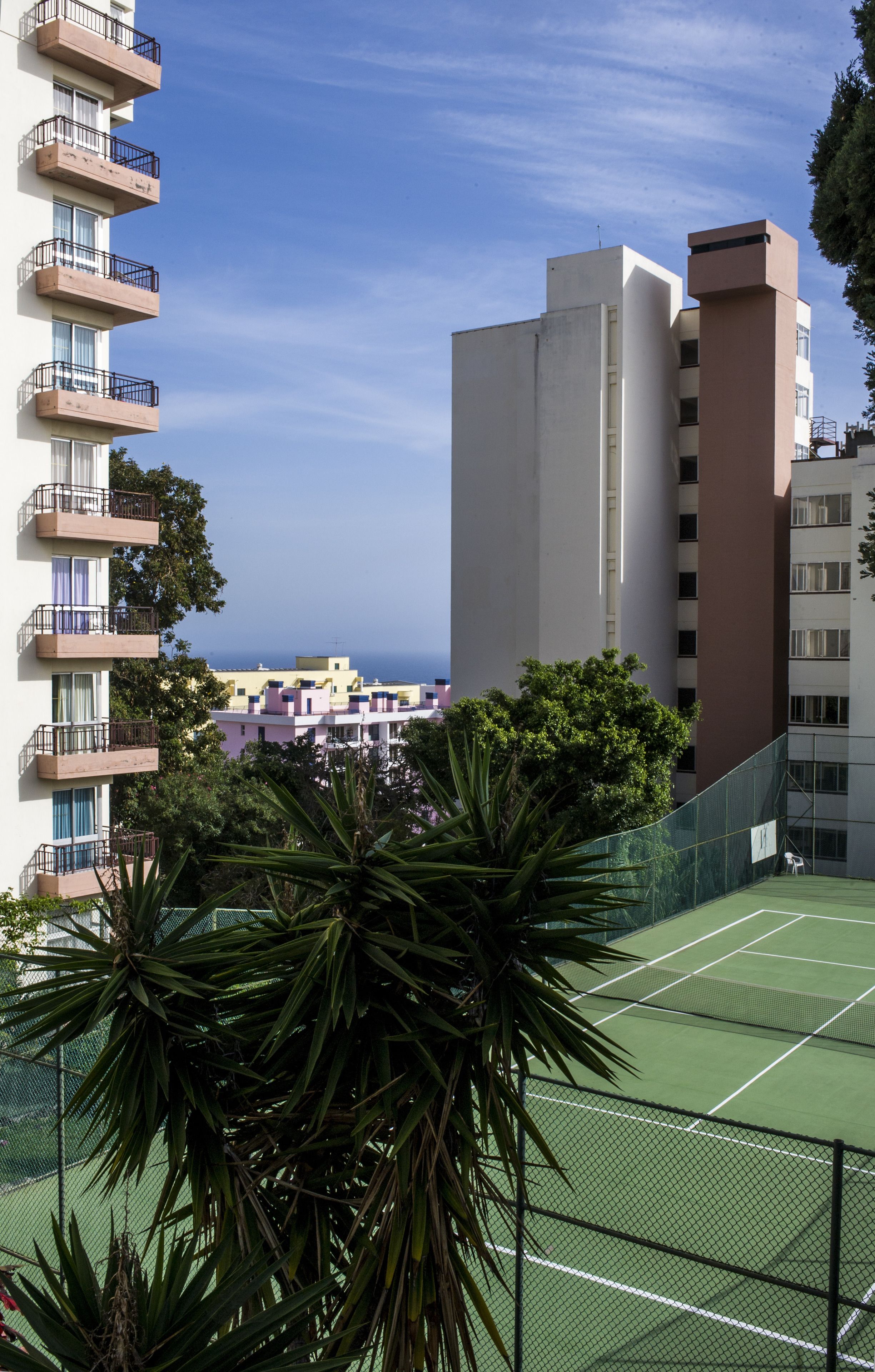 Ein Tennisplatz, umgeben von Gebäuden mit Balkonen, einem klaren Himmel und etwas Grün.