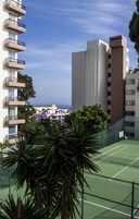 Ein Tennisplatz, umgeben von Gebäuden mit Balkonen, einem klaren Himmel und etwas Grün.
