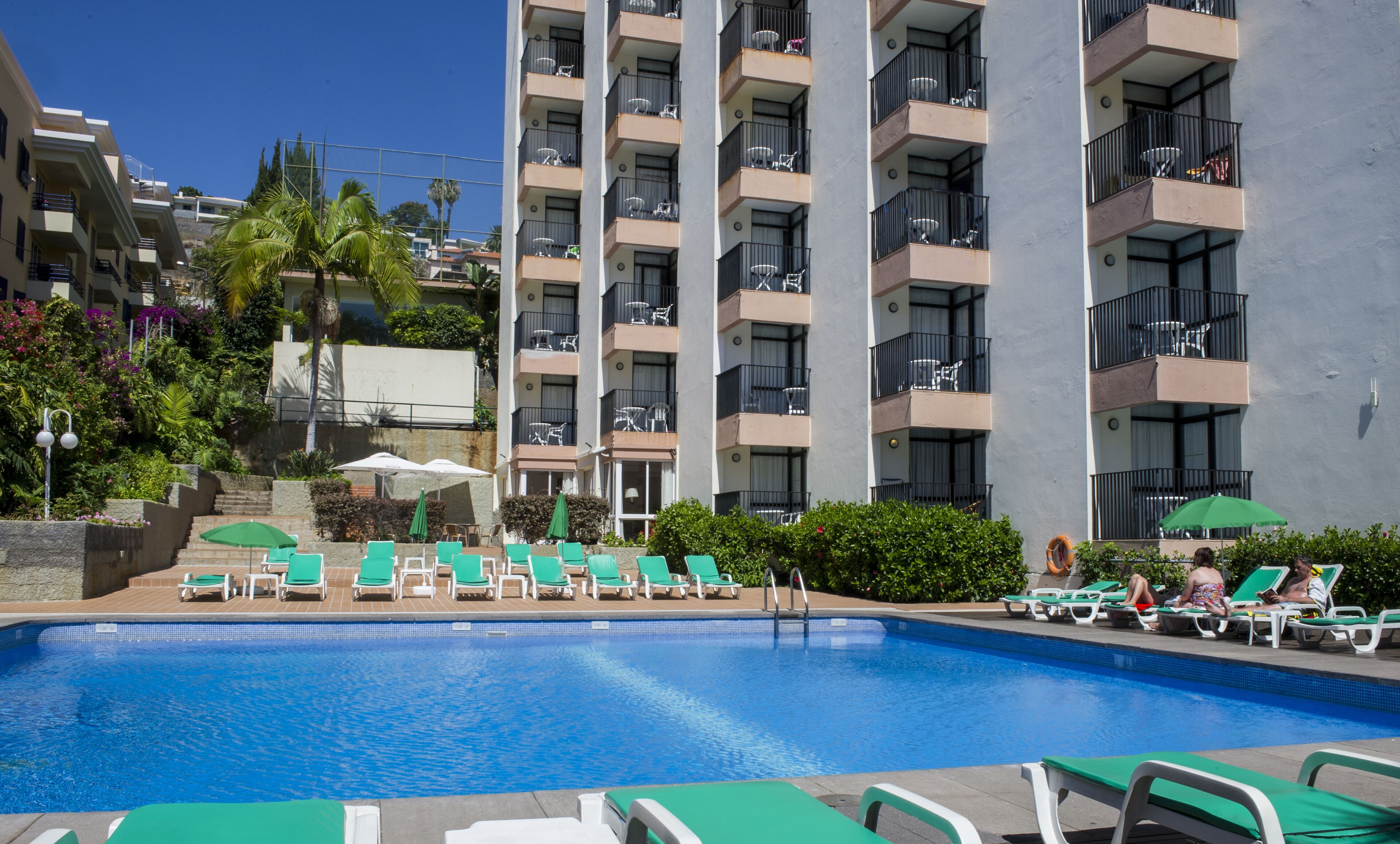A swimming pool with lounge chairs and umbrellas, surrounded by a multi-story building with balconies.