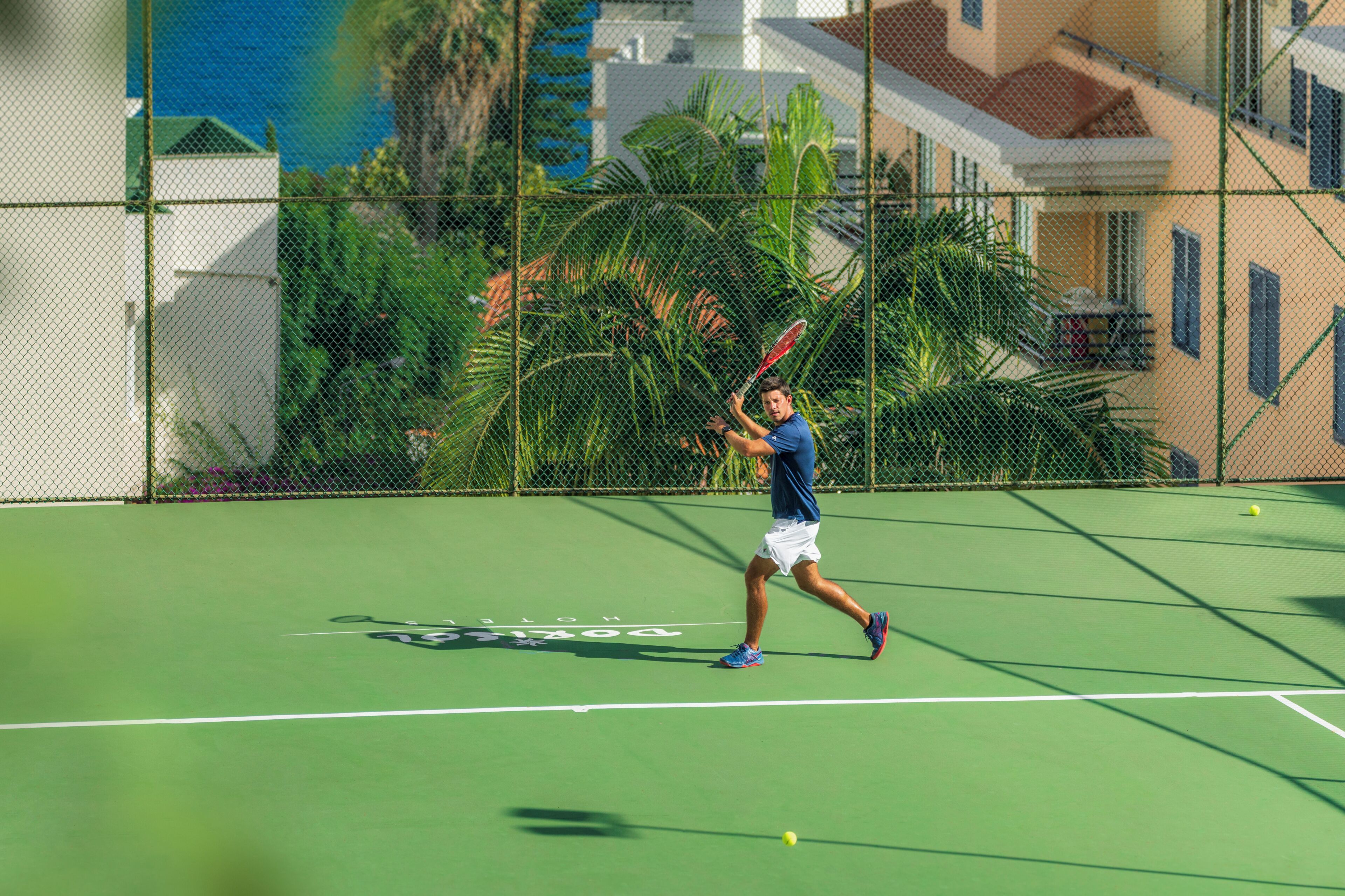 Een tennisbaan met een spelende tennisser, omringd door een hek en palmbomen.