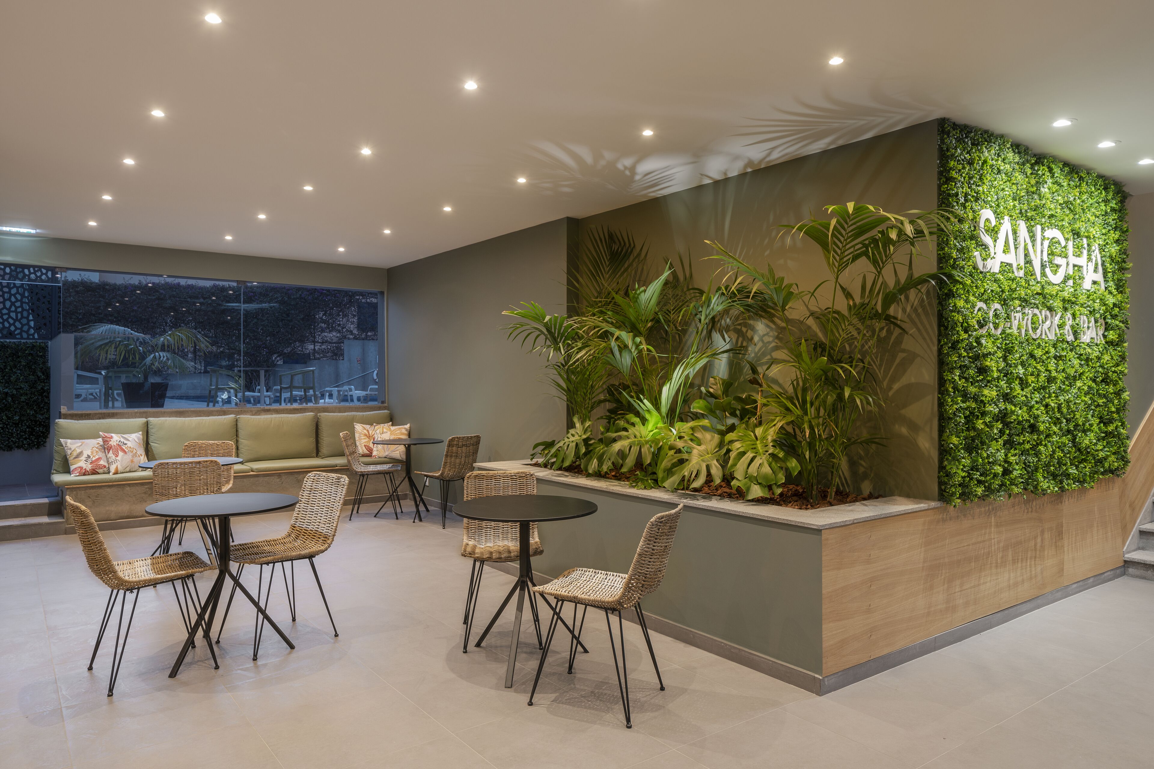 A modern lounge area with wicker furniture, a green wall with plants, and a sign reading "SANGHA CO WORKING AREA".