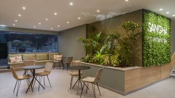 A modern lounge area with wicker furniture, a green wall with plants, and a sign reading "SANGHA CO WORKING AREA".