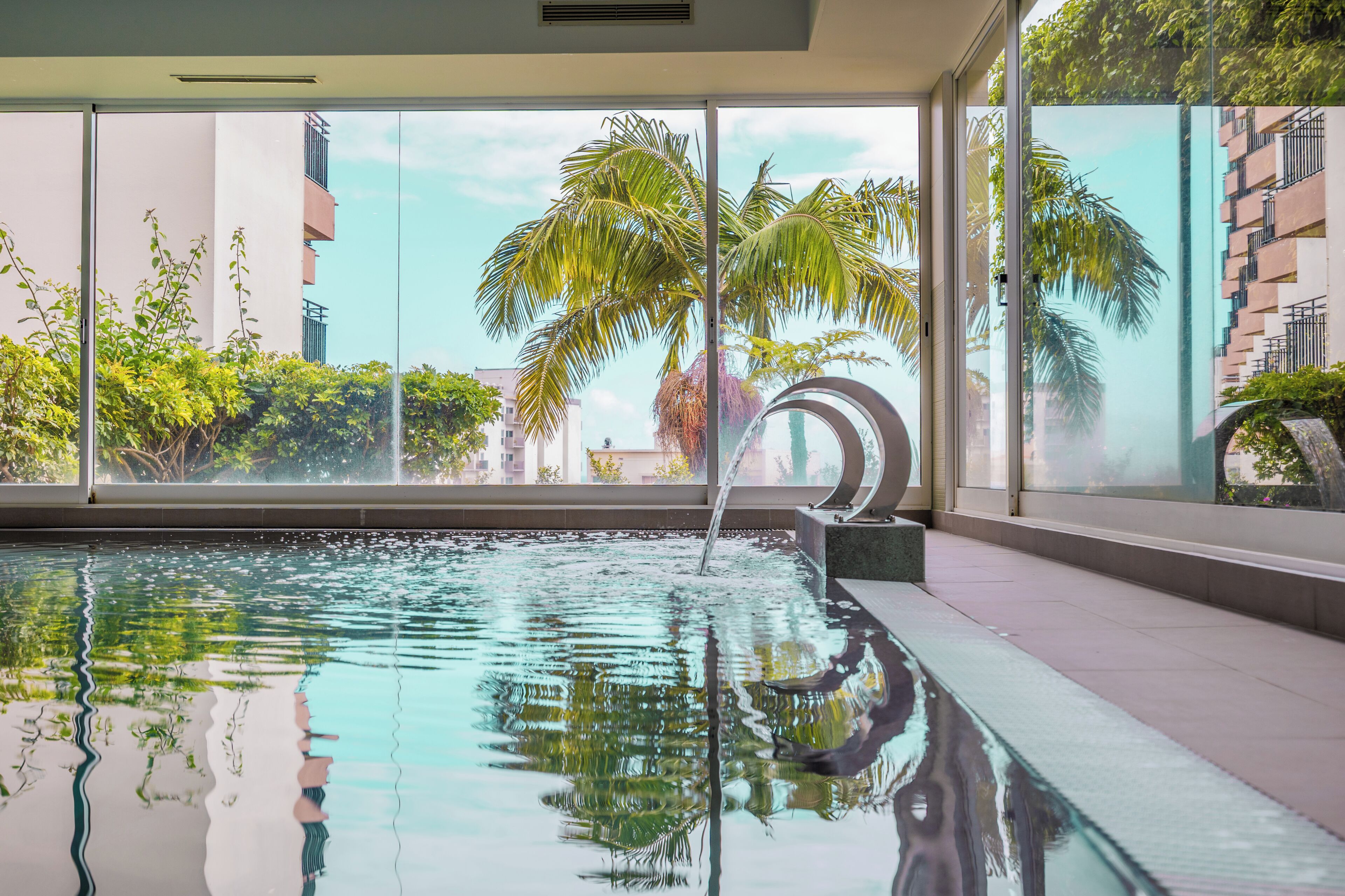 Una piscina con agua cristalina, rodeada de amplios ventanales con vistas a palmeras y edificios.