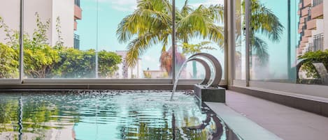 A swimming pool with clear water, surrounded by large windows offering views of palm trees and buildings.
