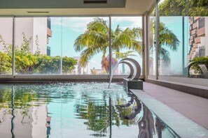 A swimming pool with clear water, surrounded by large windows offering views of palm trees and buildings. - Dorisol Buganvilia Studio Hotel (Funchal)